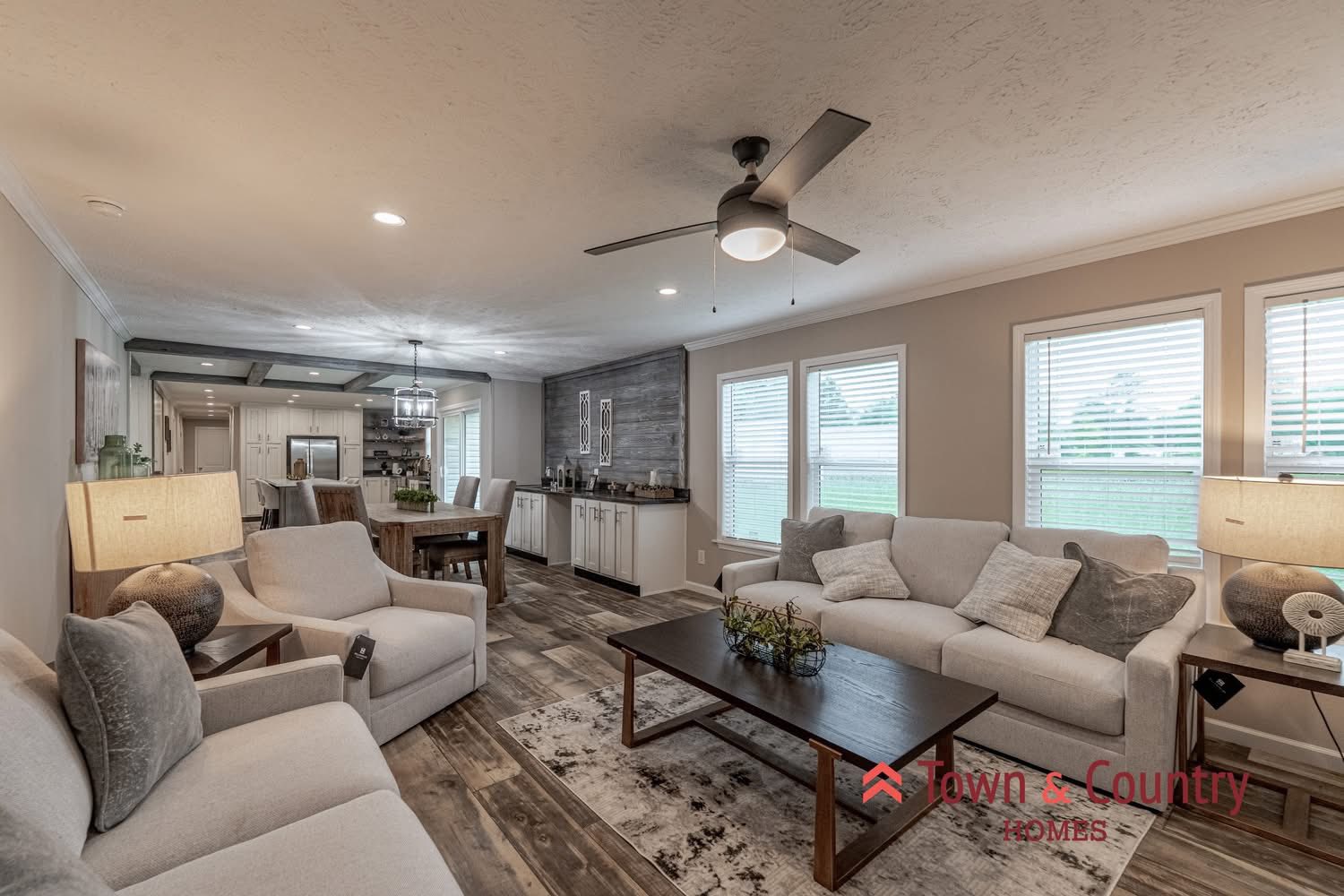 Modern living room with neutral tones, featuring two gray sofas and armchairs, a wooden coffee table, and a ceiling fan. Large windows provide natural light. The space seamlessly connects to a dining area with a rustic table and an open kitchen in the background. Cozy and inviting atmosphere.