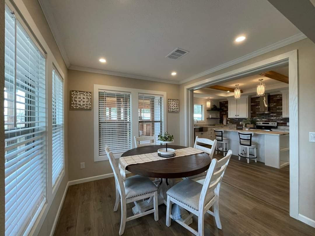 Bright dining area with a round wooden table, four white chairs, and a vase centerpiece. Kitchen view with pendant lights and modern appliances in the background.