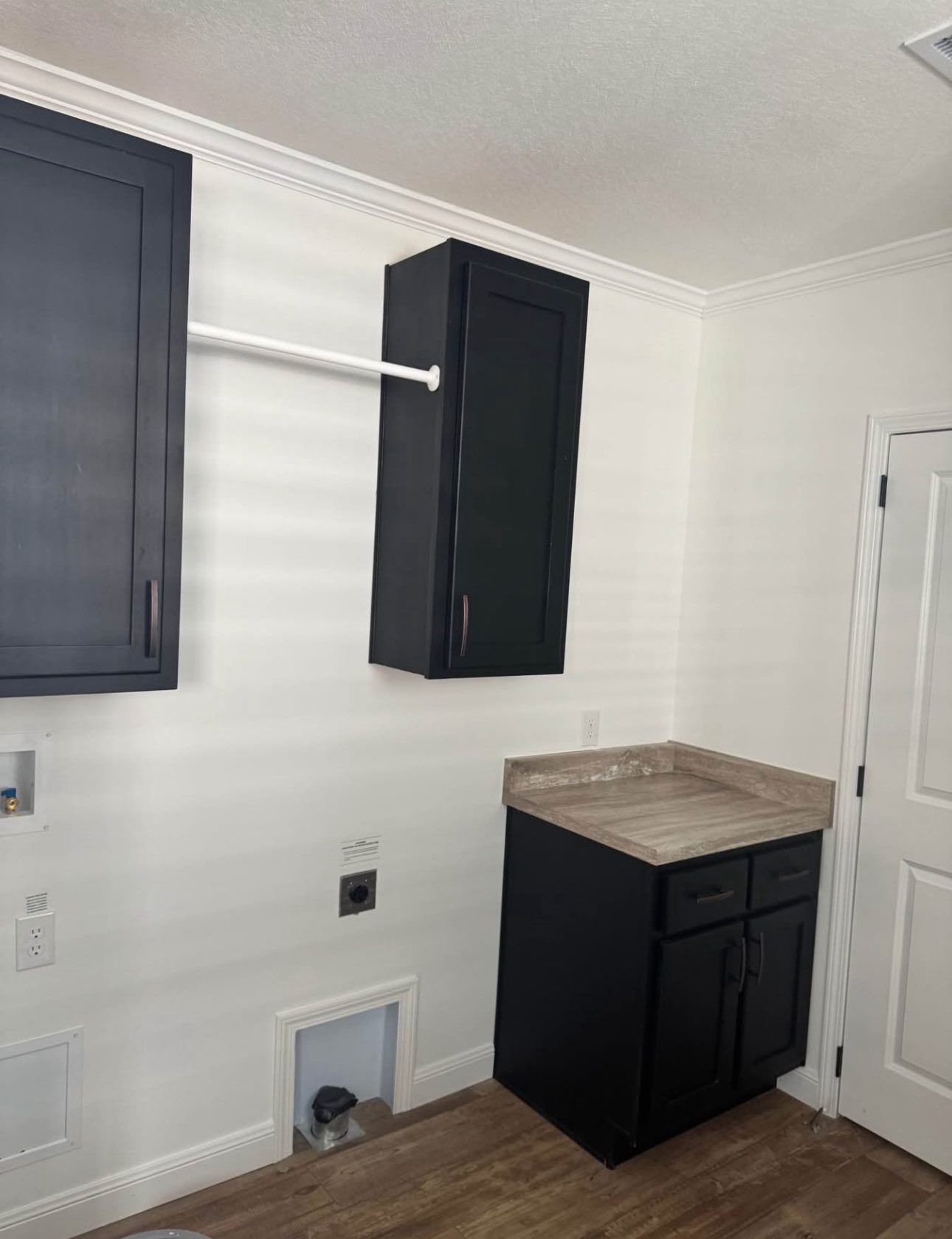 Laundry room with white walls and wood floor. Black cabinets, one overhead with a drying rod, another below with a gray countertop. Bright and clean.