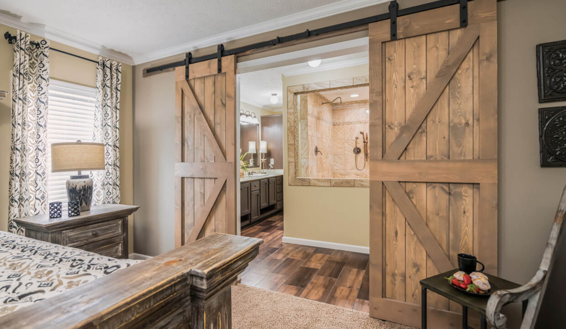 Cozy bedroom with rustic wooden barn doors leading to a bathroom. Warm tones, patterned curtains, and elegant wood flooring create a serene ambiance.