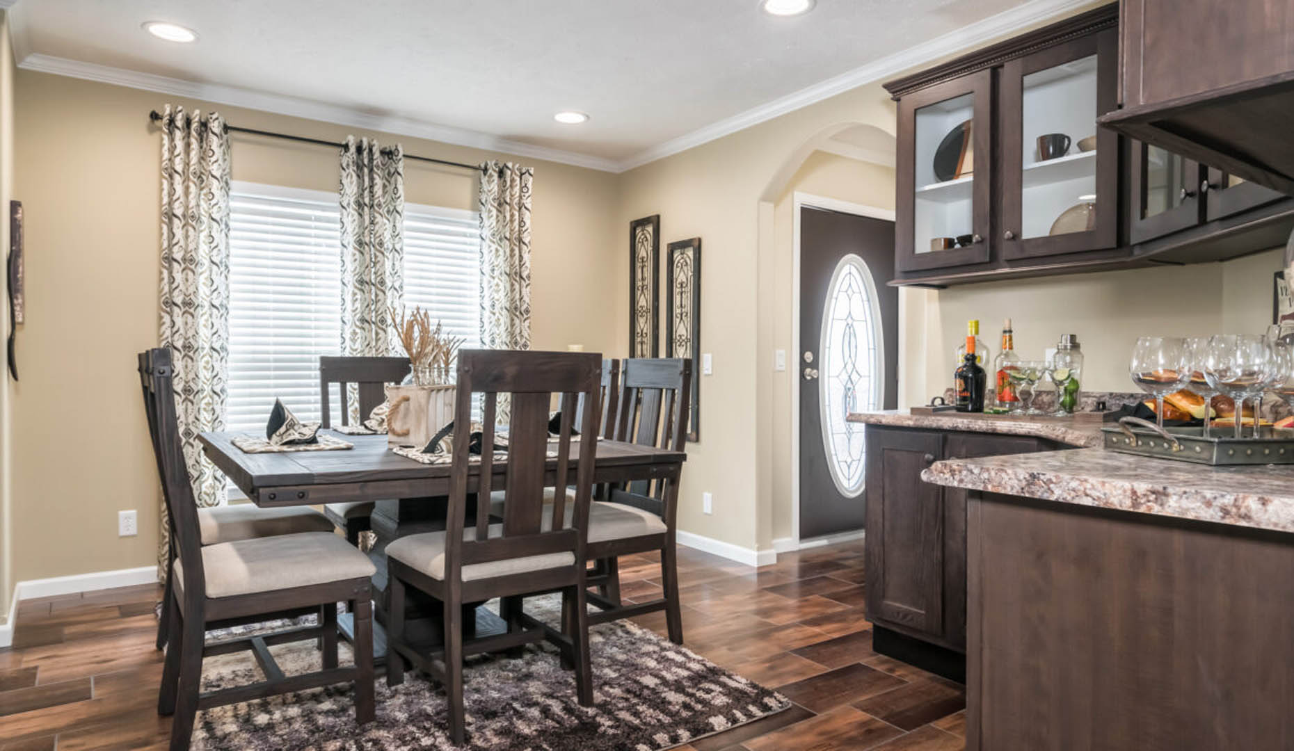 A cozy dining room with rich wooden furniture, including a table set for four. Warm lighting, patterned curtains, and a stocked bar area create a welcoming ambiance.