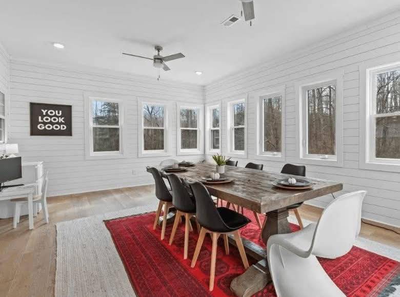 Bright dining room with white shiplap walls, a rustic wooden table, black and white chairs, and a red rug. Windows line the walls, with a sign saying "You Look Good."