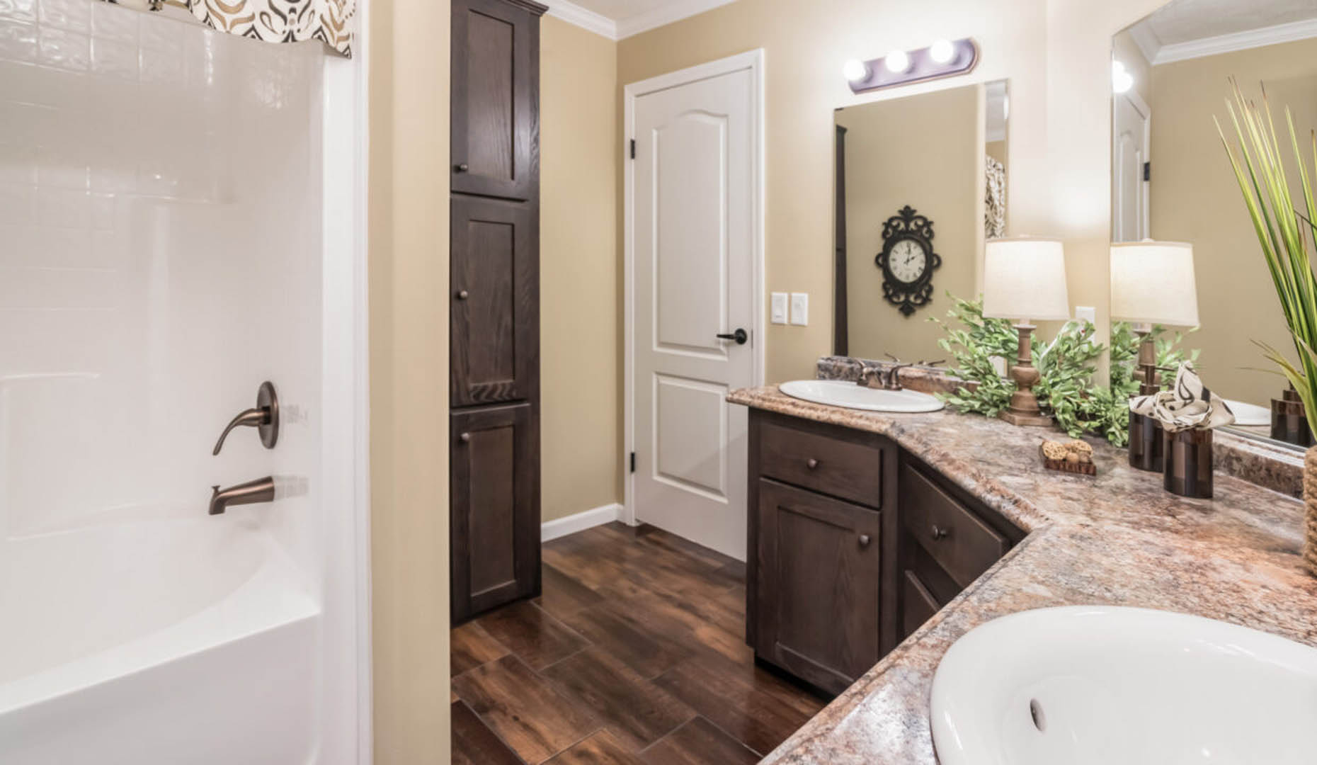 Warm-toned bathroom with wood cabinets, dual sinks, a large mirror, and plants. A bathtub is to the left. The atmosphere is cozy and inviting.