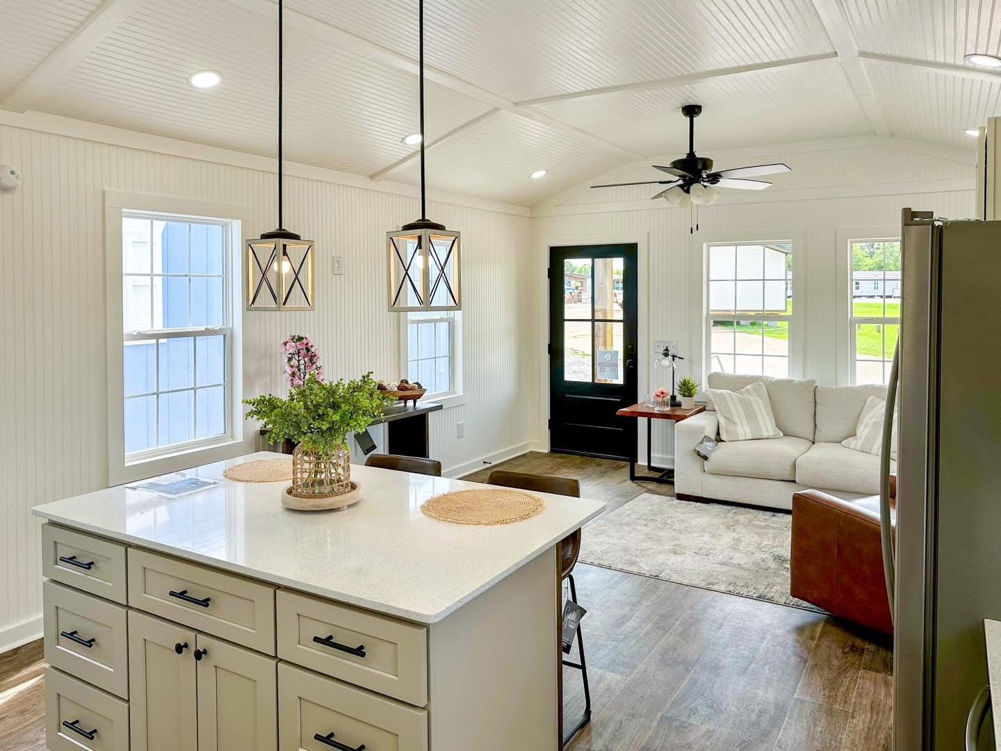 Bright open living space with white walls and vaulted ceiling. Features a kitchen island, pendant lights, sofa, and large windows, creating an airy, inviting atmosphere.