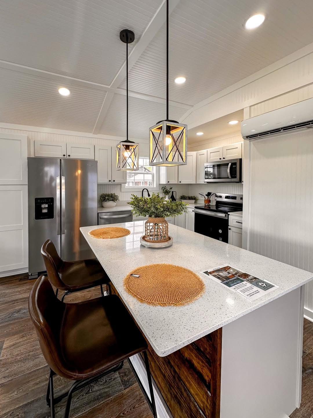 Stylish, modern kitchen with a white island, two brown chairs, geometric pendant lights, a stainless-steel fridge, and dark wood flooring. Warm, inviting tone.