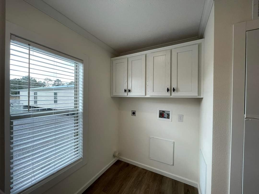 A small laundry room with white upper cabinets, a wall outlet, and plumbing connections. A window with blinds on the left lets in natural light.