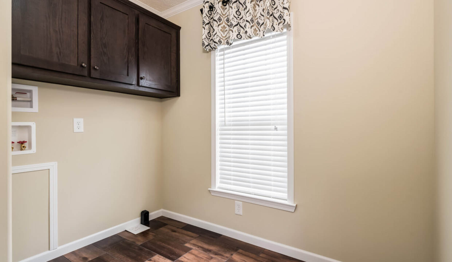 A small laundry room with beige walls and dark hardwood floors. It features dark wooden cabinets and a window with closed blinds and patterned valance.