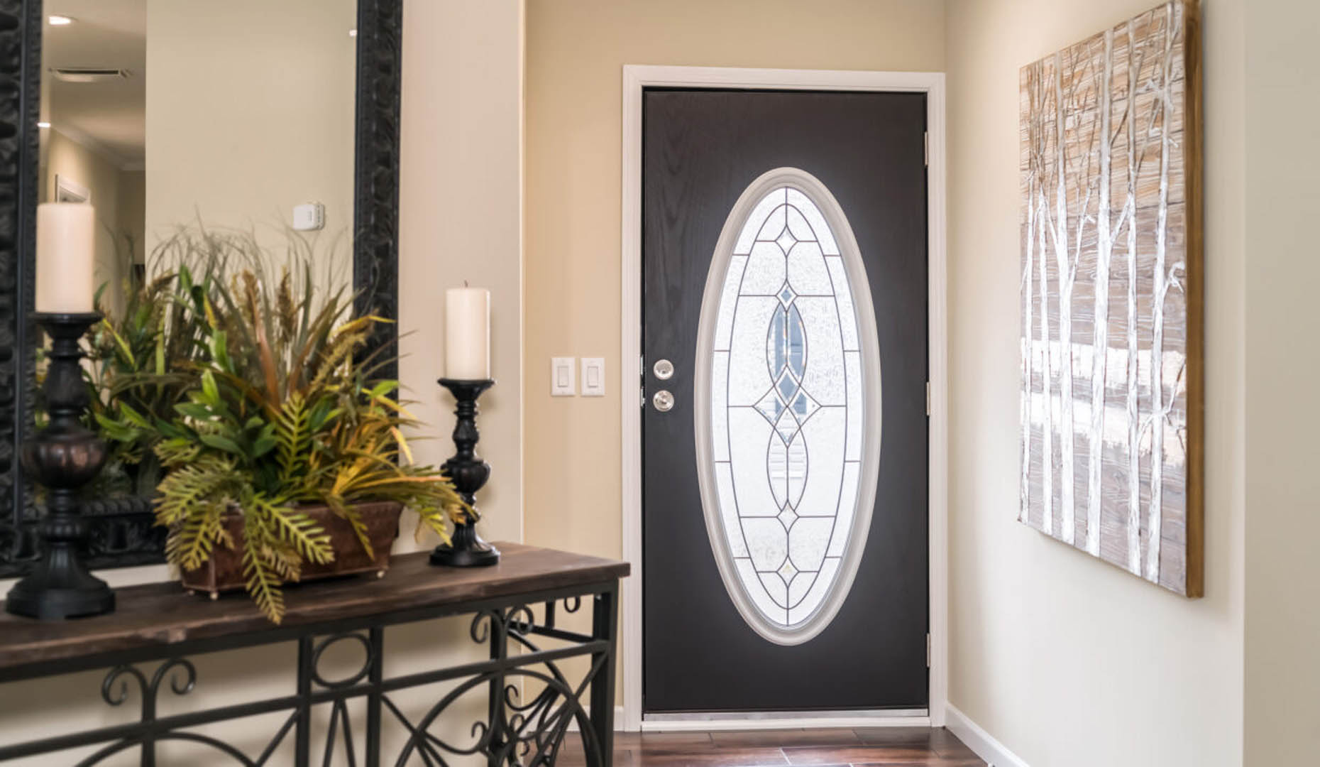 A warmly lit entryway features a dark oval-glass door, a decorative plant and candle arrangement on an ornate table, and a nature-inspired wall art piece.
