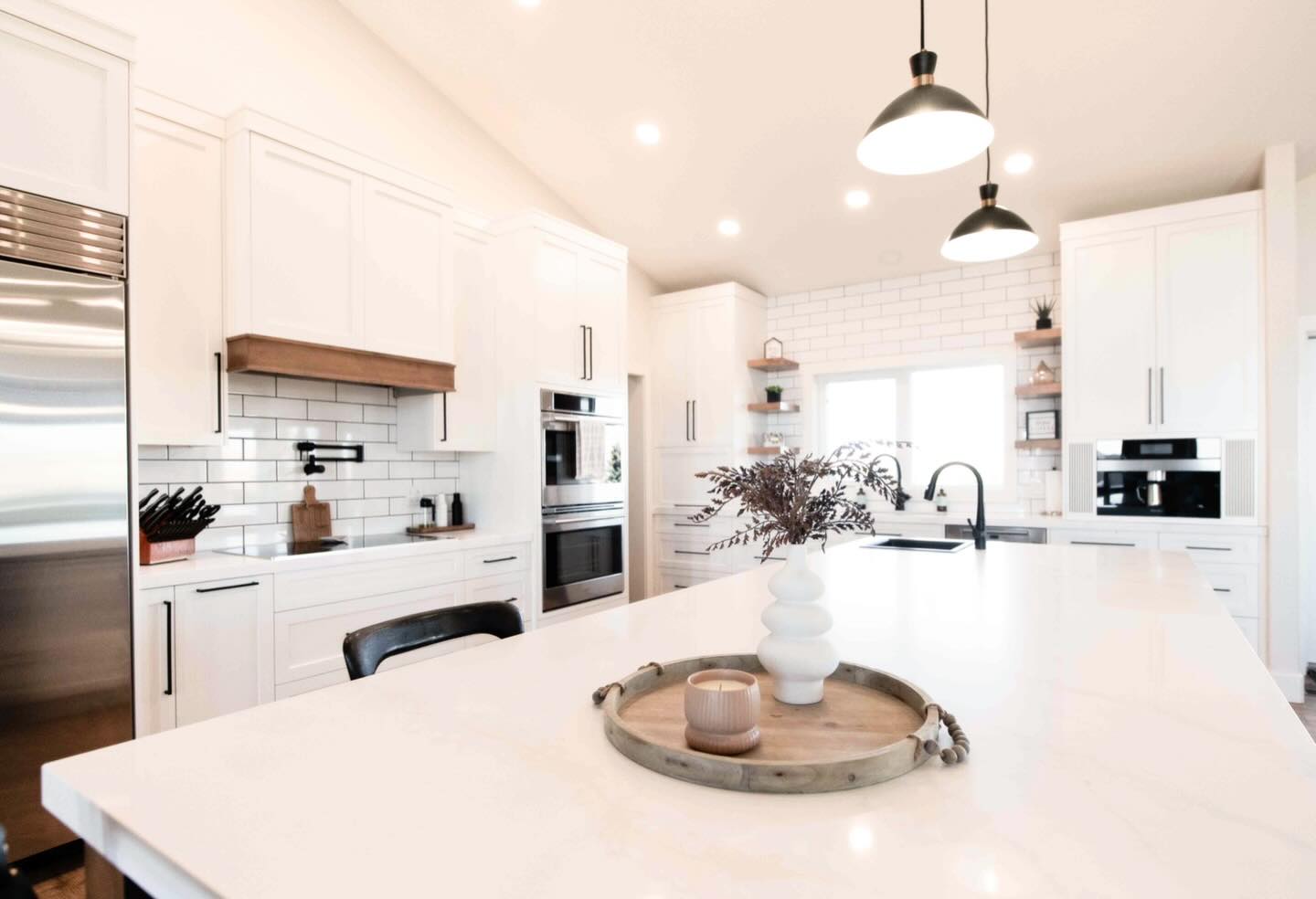 Bright, modern kitchen with white cabinets, subway tile backsplash, and a large island. Features pendant lights, a wooden tray, and minimal decor.