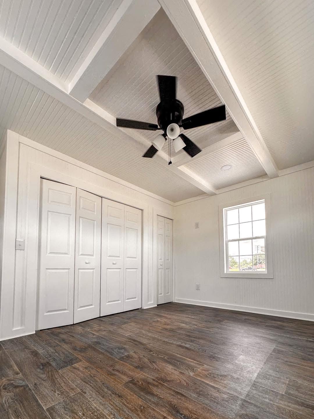 Bright minimalist room with wood floors, white paneled walls, and a vaulted ceiling. A ceiling fan is centered, and closets with white doors line one wall.