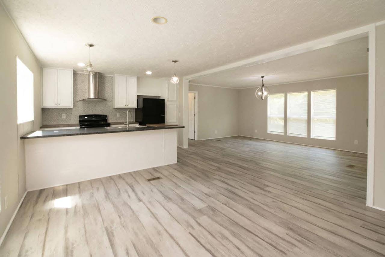 Spacious open-concept room with light wood flooring, white kitchen cabinets, black countertops, modern appliances, and large windows, creating an airy feel.