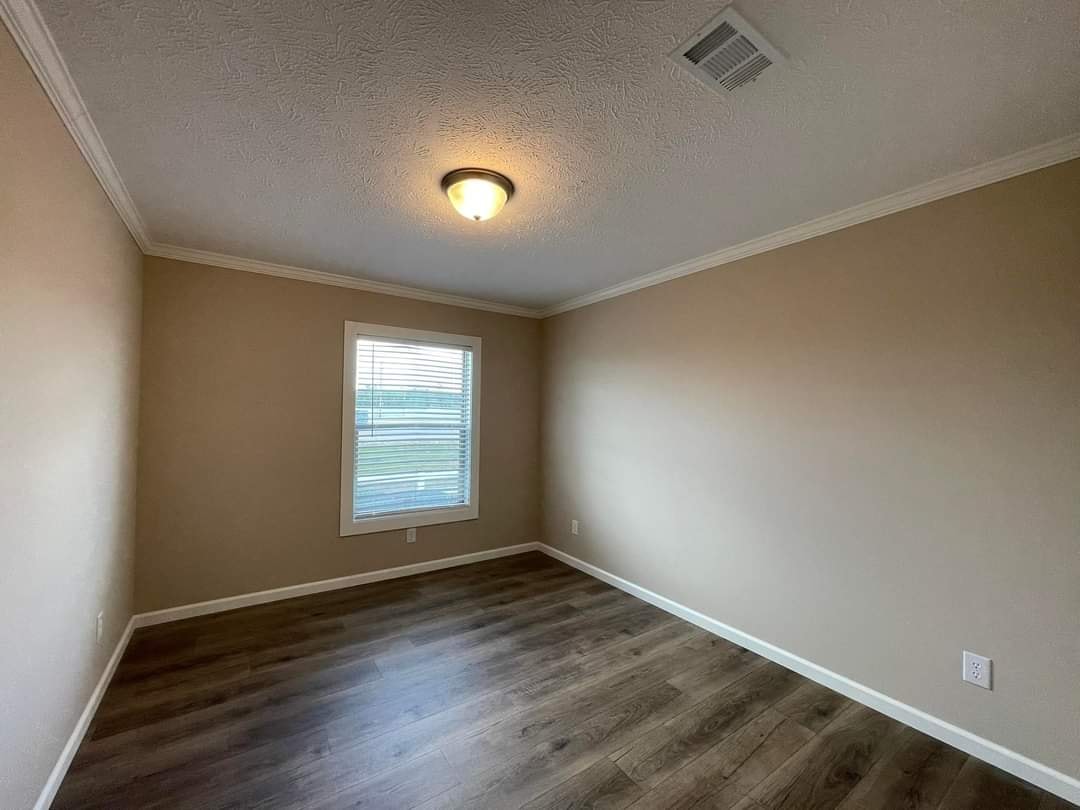 A small, empty room with beige walls and wood flooring. A window with blinds on the left wall lets in natural light. A ceiling light fixture is lit.
