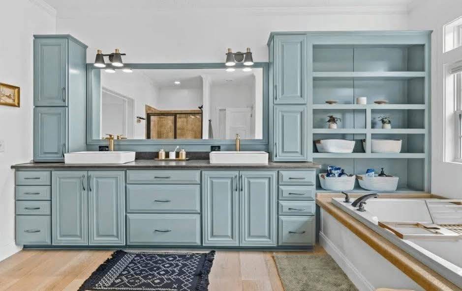 Elegant bathroom with blue cabinetry featuring twin sinks, large mirror, and open shelving. Soft lighting creates a calm, sophisticated ambiance.