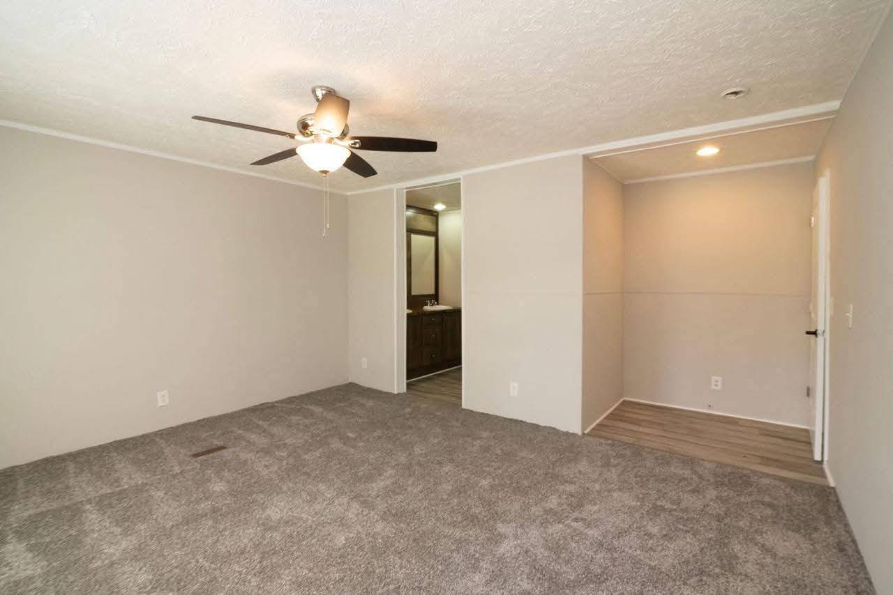 Minimalist room with gray carpet, beige walls, and a ceiling fan with lights. An open door leads to a bathroom with dark wood cabinets, creating a neutral, calm tone.