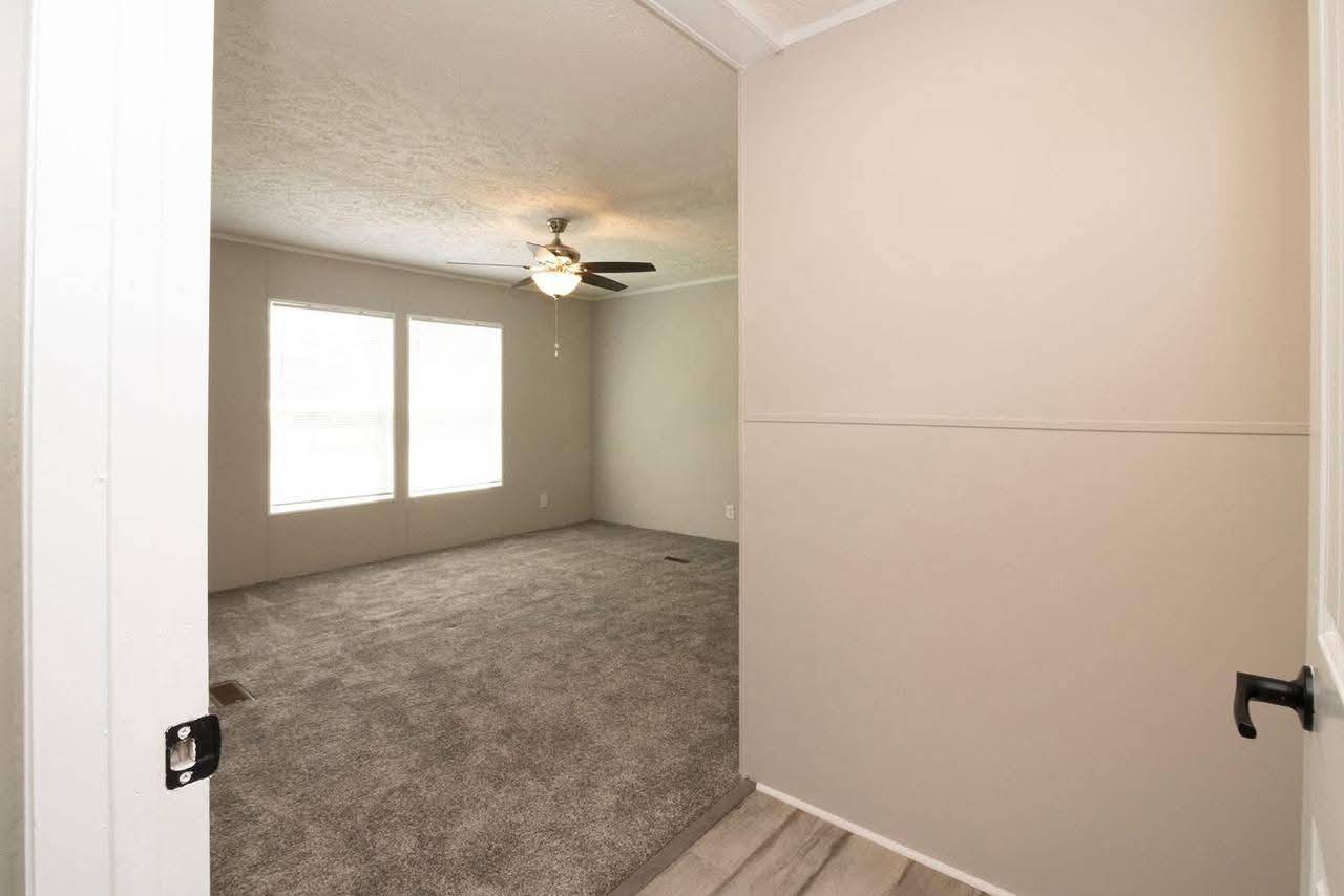 Empty room with beige walls, a ceiling fan, and two large windows on one side. The floor has neutral-toned carpet, creating a calm and spacious feel.