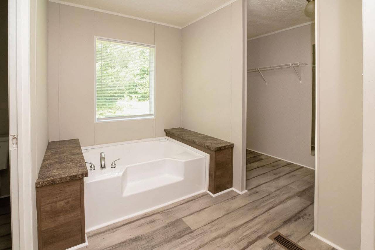Modern bathroom with a built-in white bathtub centered under a window. Brown countertops and wood-like flooring create a calm, minimalist atmosphere.