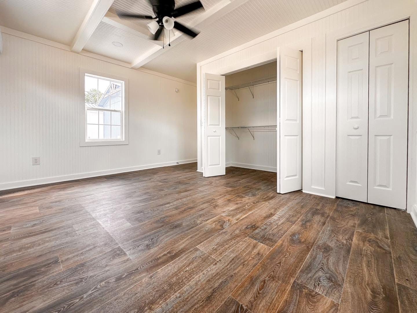 Empty room with wood-look flooring, white walls, and beamed ceiling. Features a ceiling fan, a window, and an open closet with shelving. Calm ambiance.