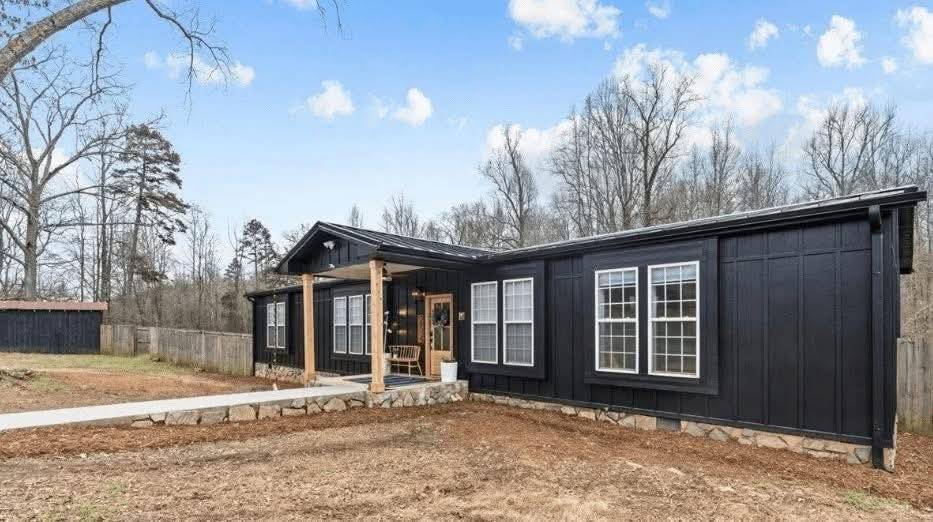 Single-story black house with white-trimmed windows, a wooden porch, and a stone-lined base, set in a grassy yard with bare trees under a blue sky.