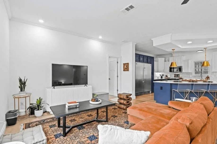 Modern living room with a rust-colored sectional, large TV, and blue-accented kitchen. Warm lighting, houseplants, and patterned rug create a cozy atmosphere.