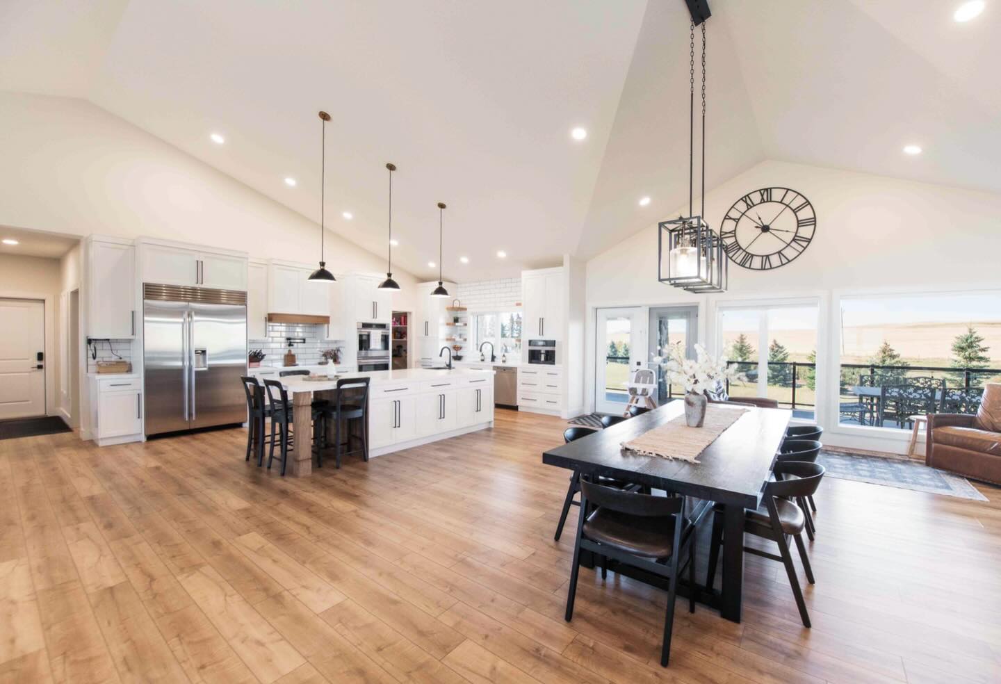 Spacious modern kitchen and dining area with white cabinets, wooden floors, and pendant lights. Large windows offer natural light and outdoor views.