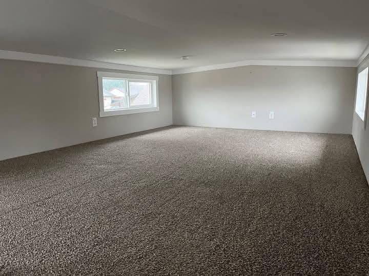 A small, low-ceilinged room with beige carpet and light gray walls. Two rectangular windows bring light, creating a cozy, compact space.