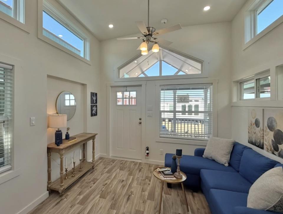 Bright living room with a high ceiling, white walls, and light wood flooring. Features a cozy blue sofa, wooden side table, lamp, and circular mirror.