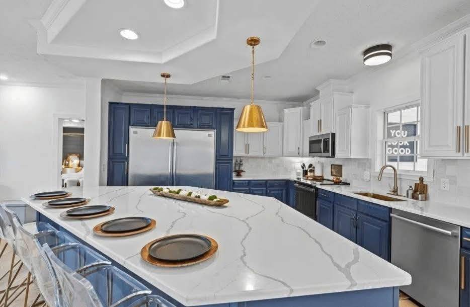 Elegant kitchen featuring a large marble island with six black and gold plates, gold pendant lights, navy and white cabinetry, and stainless steel appliances.