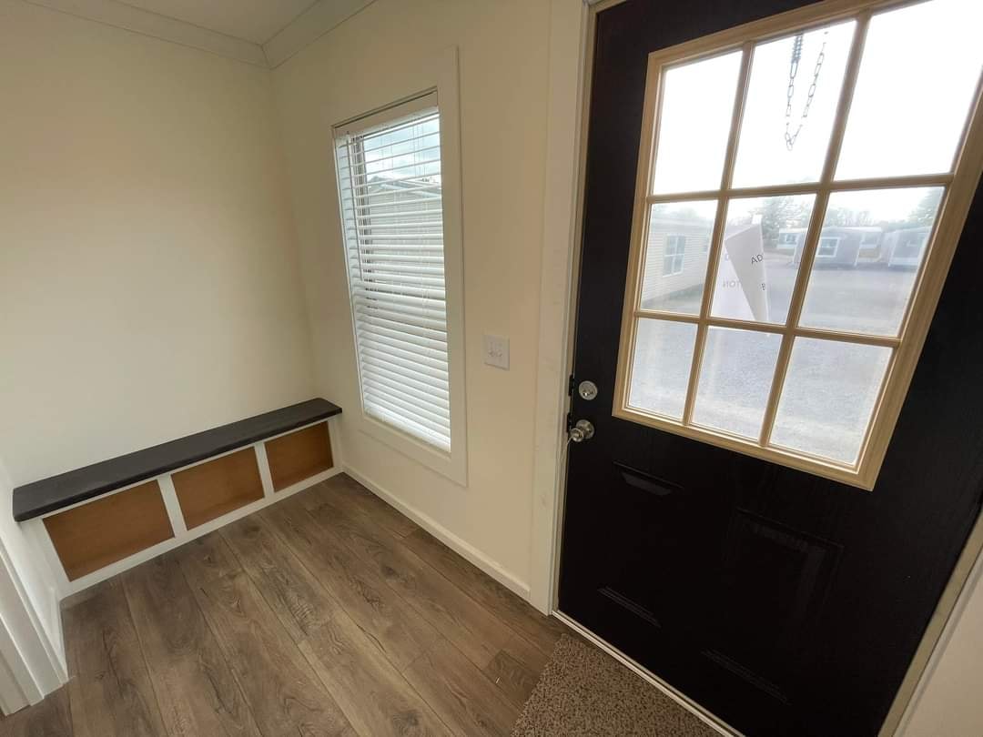 Small entryway with a dark wood door featuring glass panes. A window with blinds is on the left, above a bench with storage. Hardwood floor adds warmth.