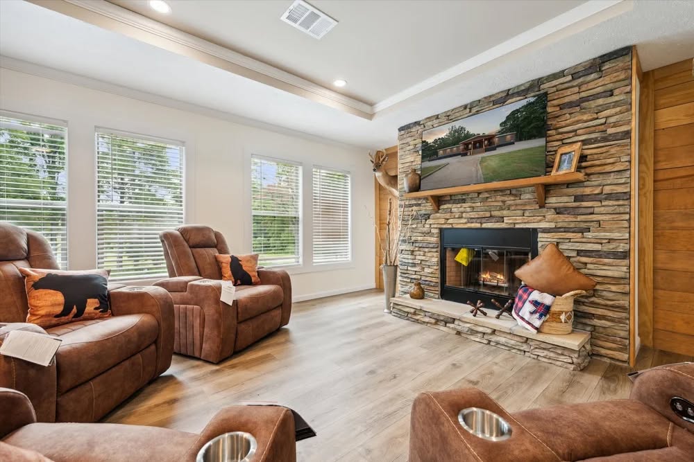 Cozy living room with brown reclining chairs and bear-themed pillows. A stone fireplace features a large photo above and shelves with décor. Light streams through large windows, highlighting the warm, inviting atmosphere.