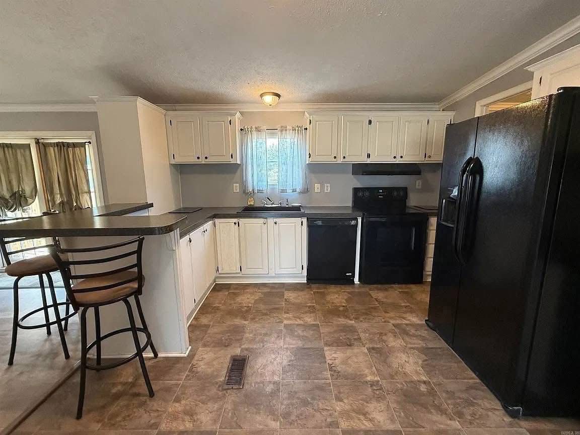 A modern kitchen with white cabinets, black appliances, and a large black fridge. There's a central light, window with curtains, and two bar stools. The flooring is tiled, creating a cozy ambiance.