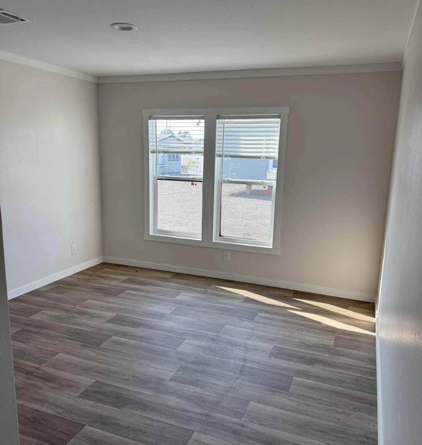 An empty room with light gray walls, wood laminate flooring, and two rectangular windows with partially open blinds letting in natural light.