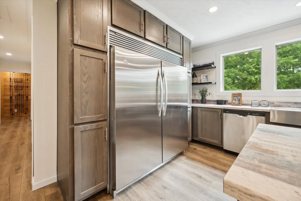 Modern kitchen with a large stainless steel fridge, wooden cabinetry, and light wood flooring. Bright natural light from windows creates an inviting atmosphere.