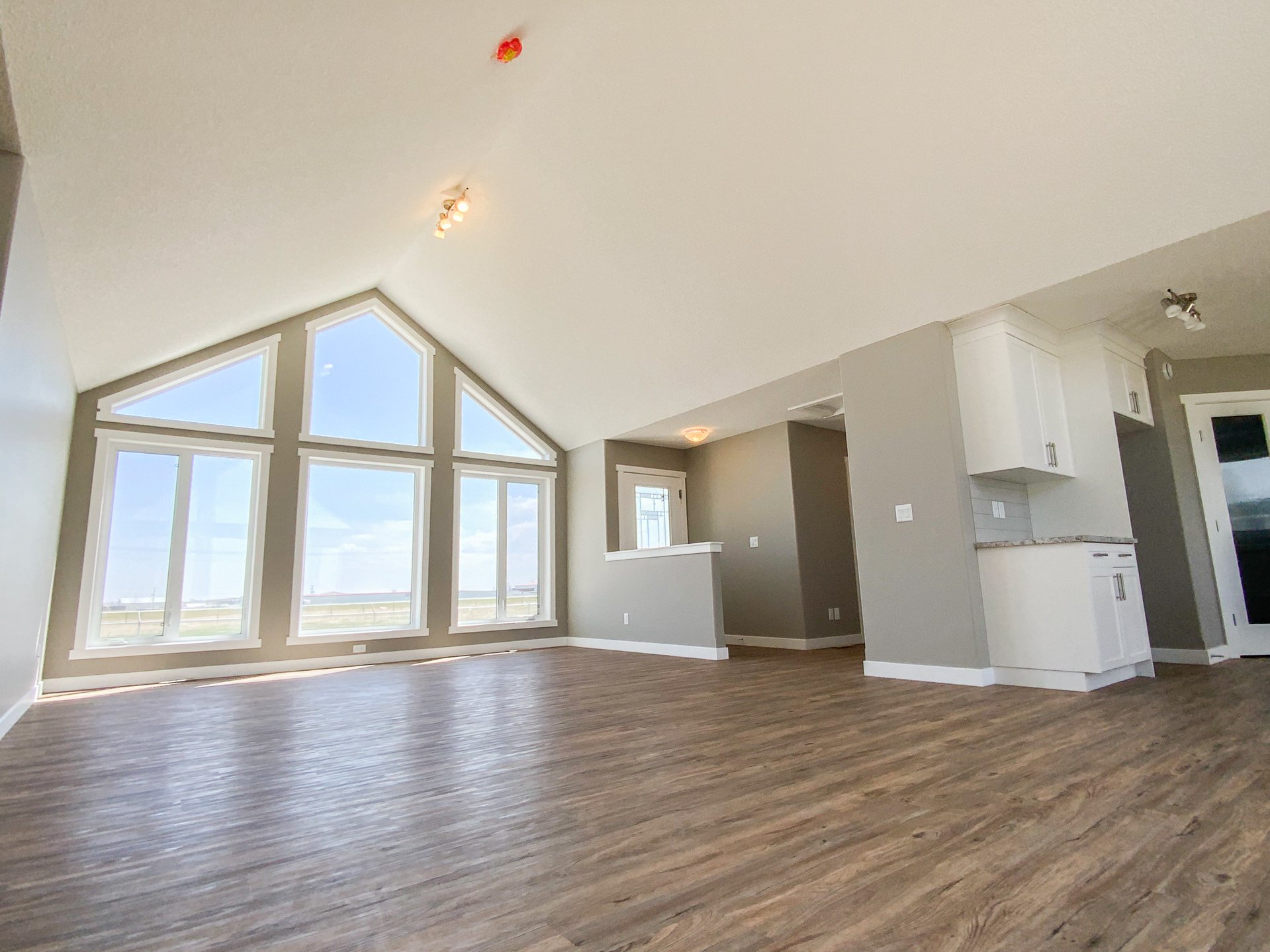 Spacious, bright living room with large triangular windows revealing a clear sky. Features light hardwood floors and a view of a modern kitchen.