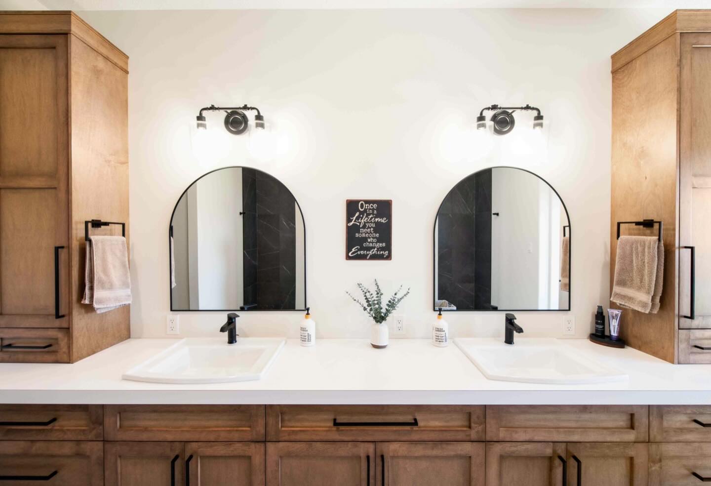 Modern bathroom with double sinks, wooden cabinets, and arched mirrors. Bright lights and a small plant create a fresh, inviting atmosphere.