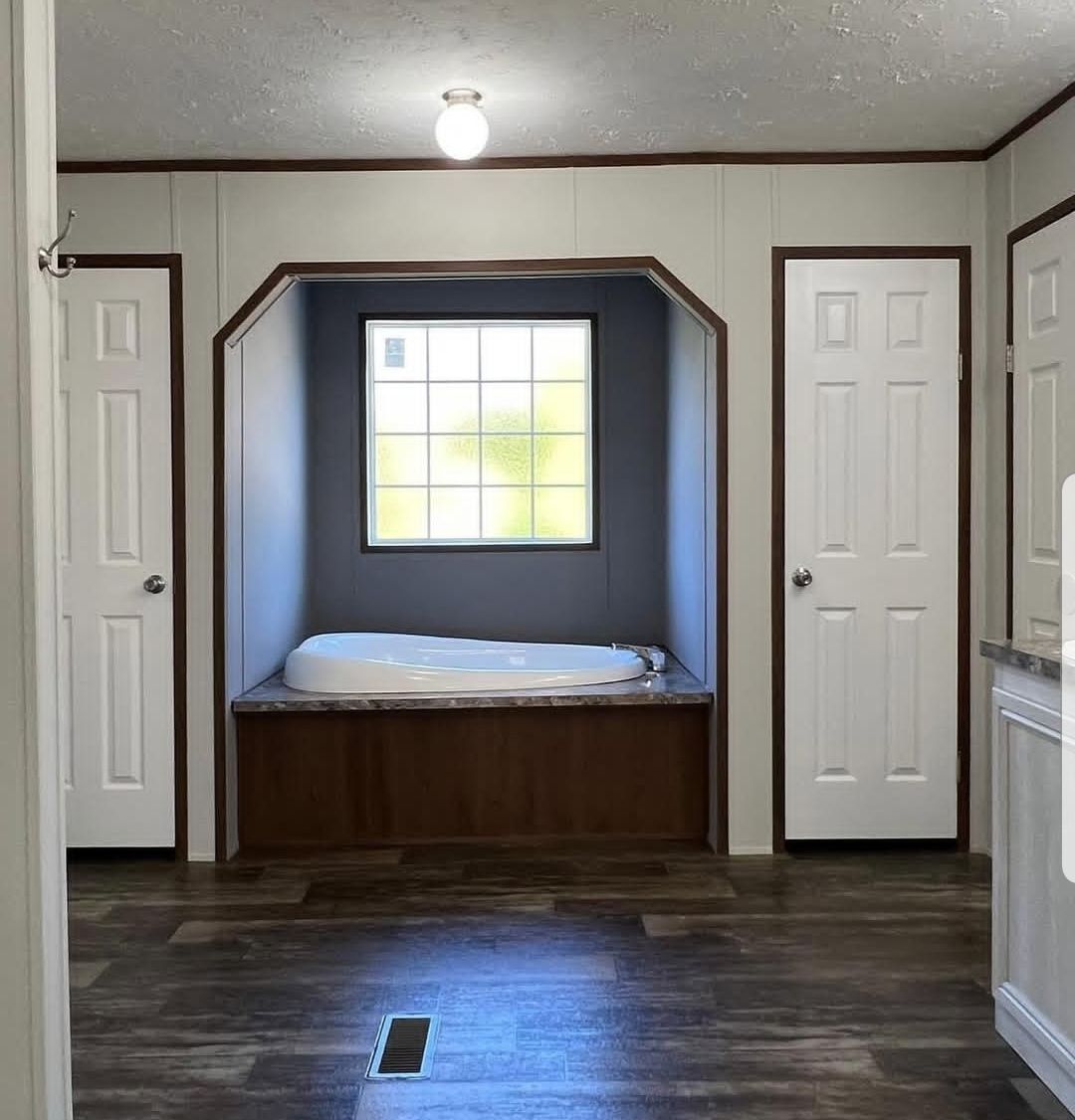 Simple bathroom with a white tub set into a wooden alcove under a grid window. Two white doors flank the tub, and hardwood flooring adds warmth.