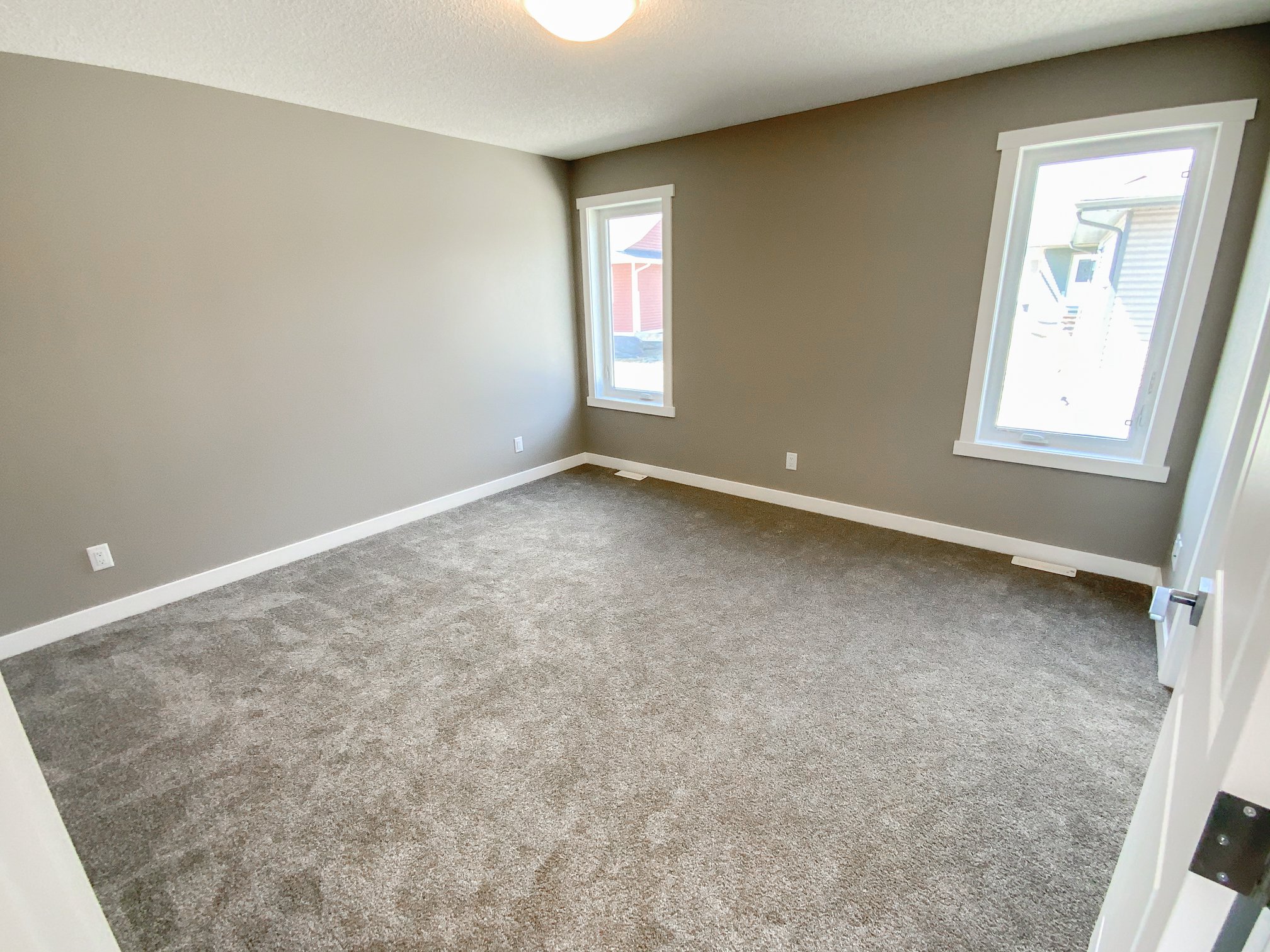 An empty room with beige carpet, light gray walls, and two windows with white trim. The space is bright with natural light, conveying a clean, open feel.