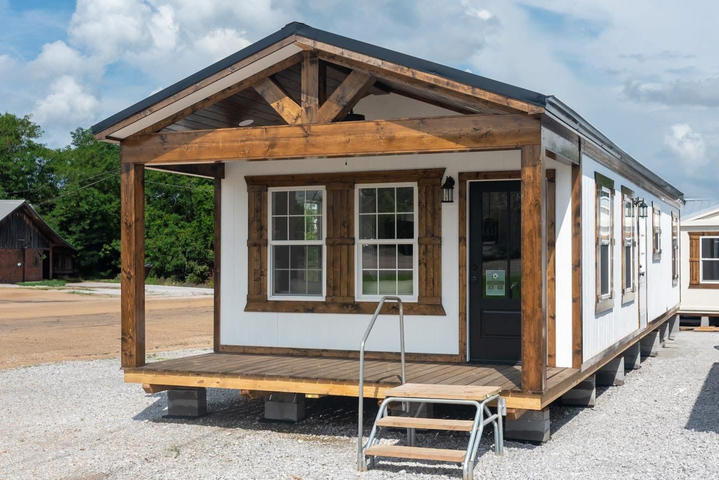A small, white manufactured home with wooden trim and a gable roof sits on a gravel lot. It has a front porch with metal steps and a cloudy sky backdrop.