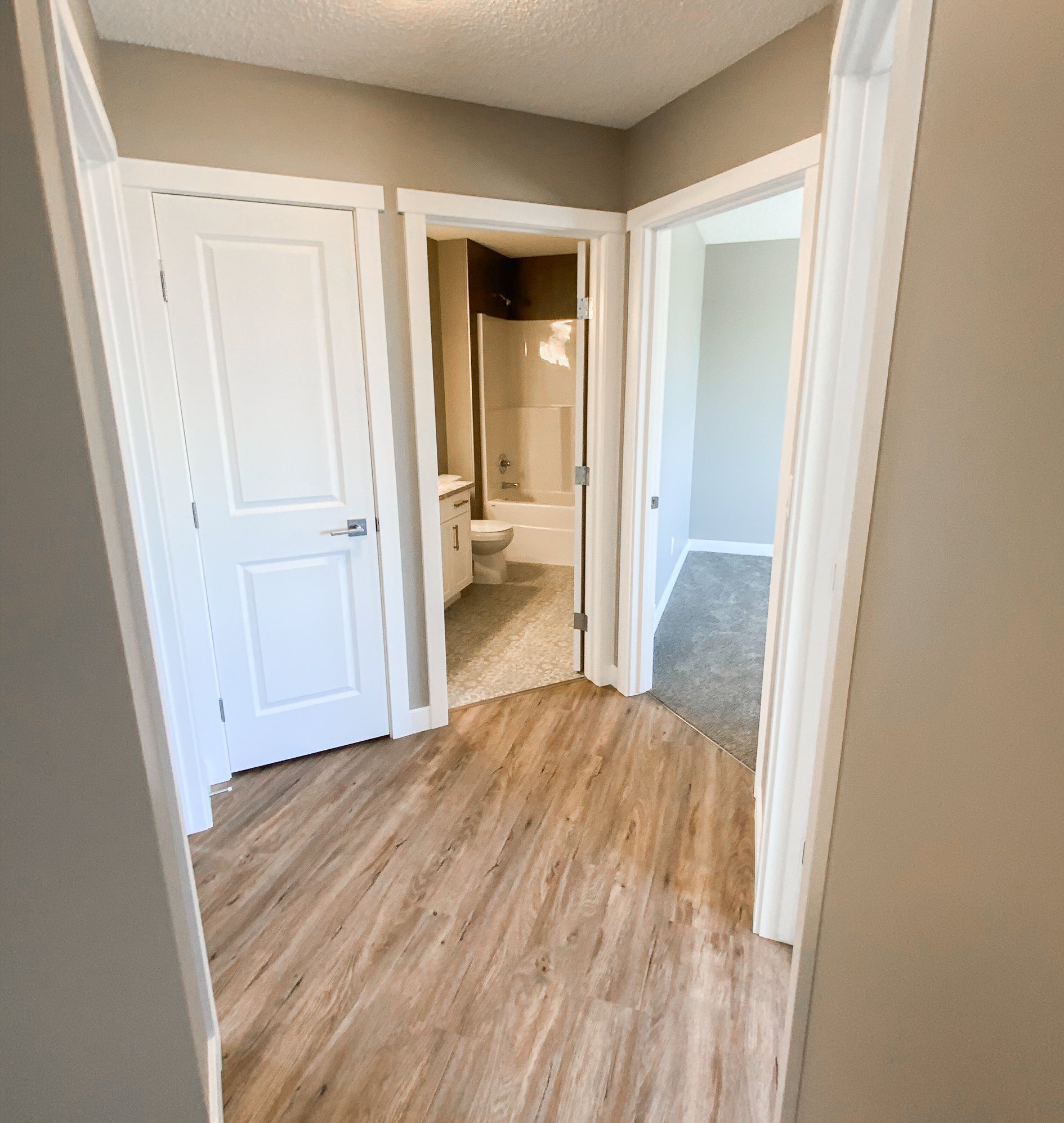 Hallway with light wood flooring, leading to three doors. A bathroom is visible straight ahead, with neutral walls creating a calm atmosphere.