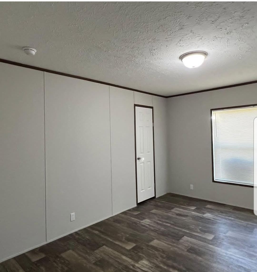 An empty room with gray walls, dark wood flooring, and a white ceiling light. A door and a window with closed blinds are on adjacent walls. Minimalist and clean.