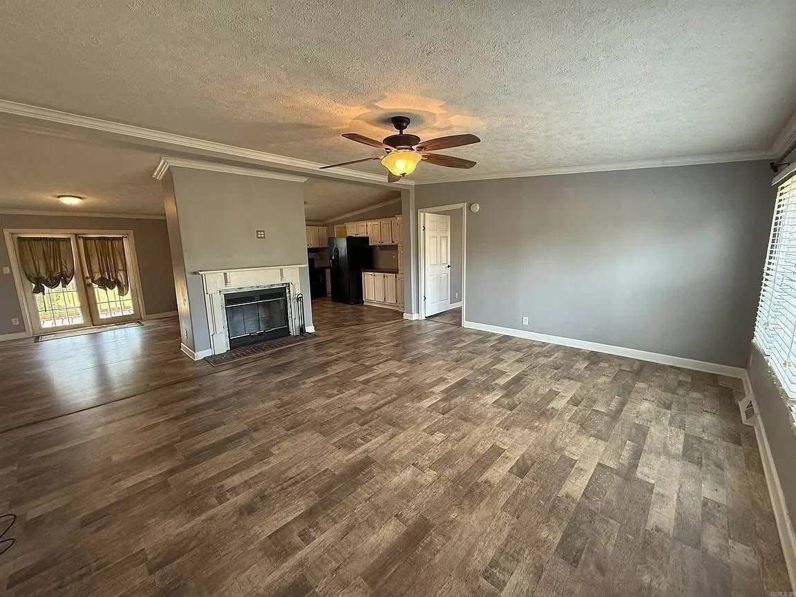 Spacious living room with wooden flooring, a ceiling fan, and neutral gray walls. A fireplace adds coziness, adjacent to the open kitchen.