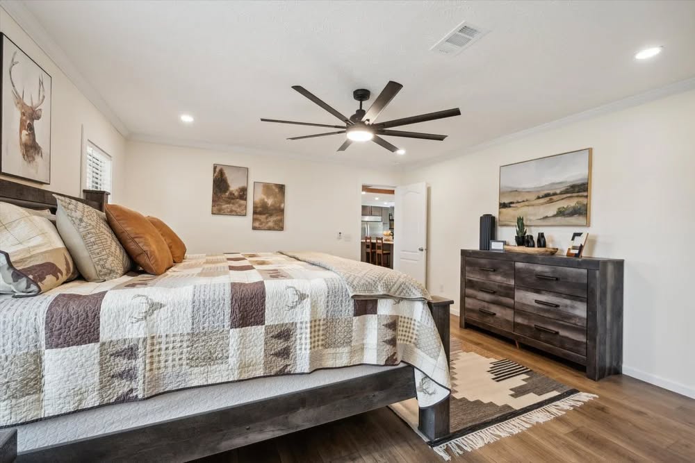 Spacious bedroom with a rustic theme. Features a large bed with a patchwork quilt, wooden dresser, and nature-themed artwork. Warm, cozy ambiance.