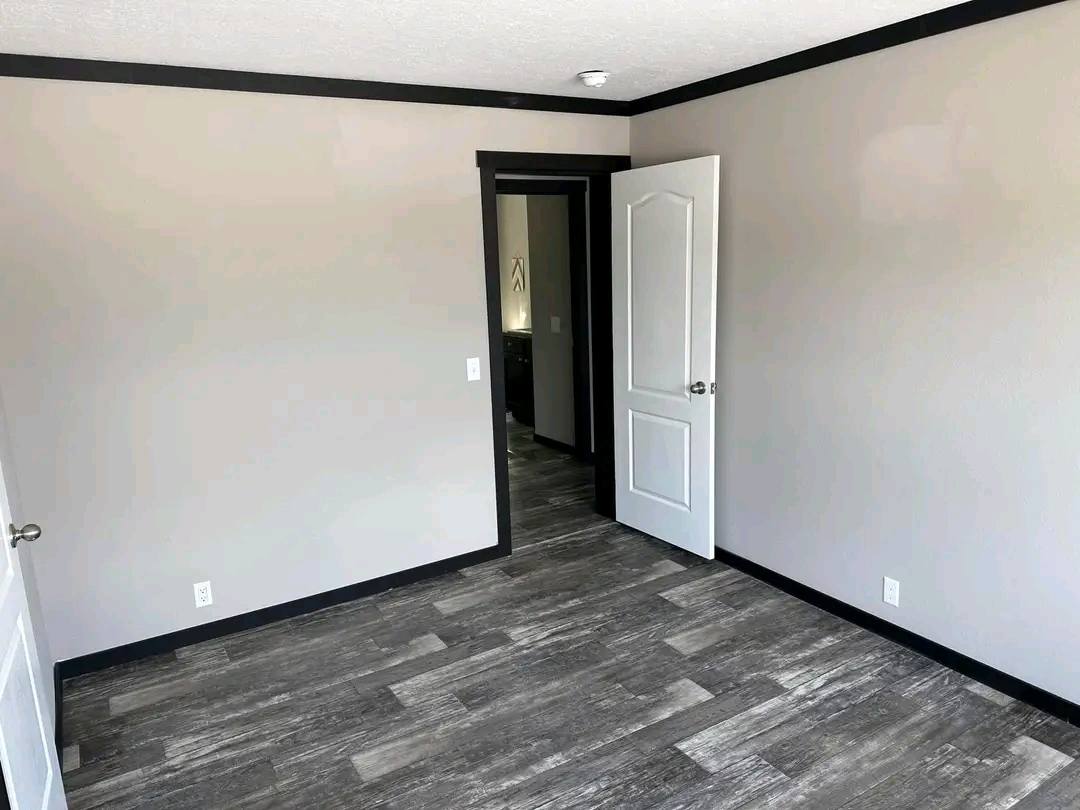 An empty room with light gray walls and dark trim. It features a wooden floor and an open door leading to a hallway, creating a clean, modern feel.