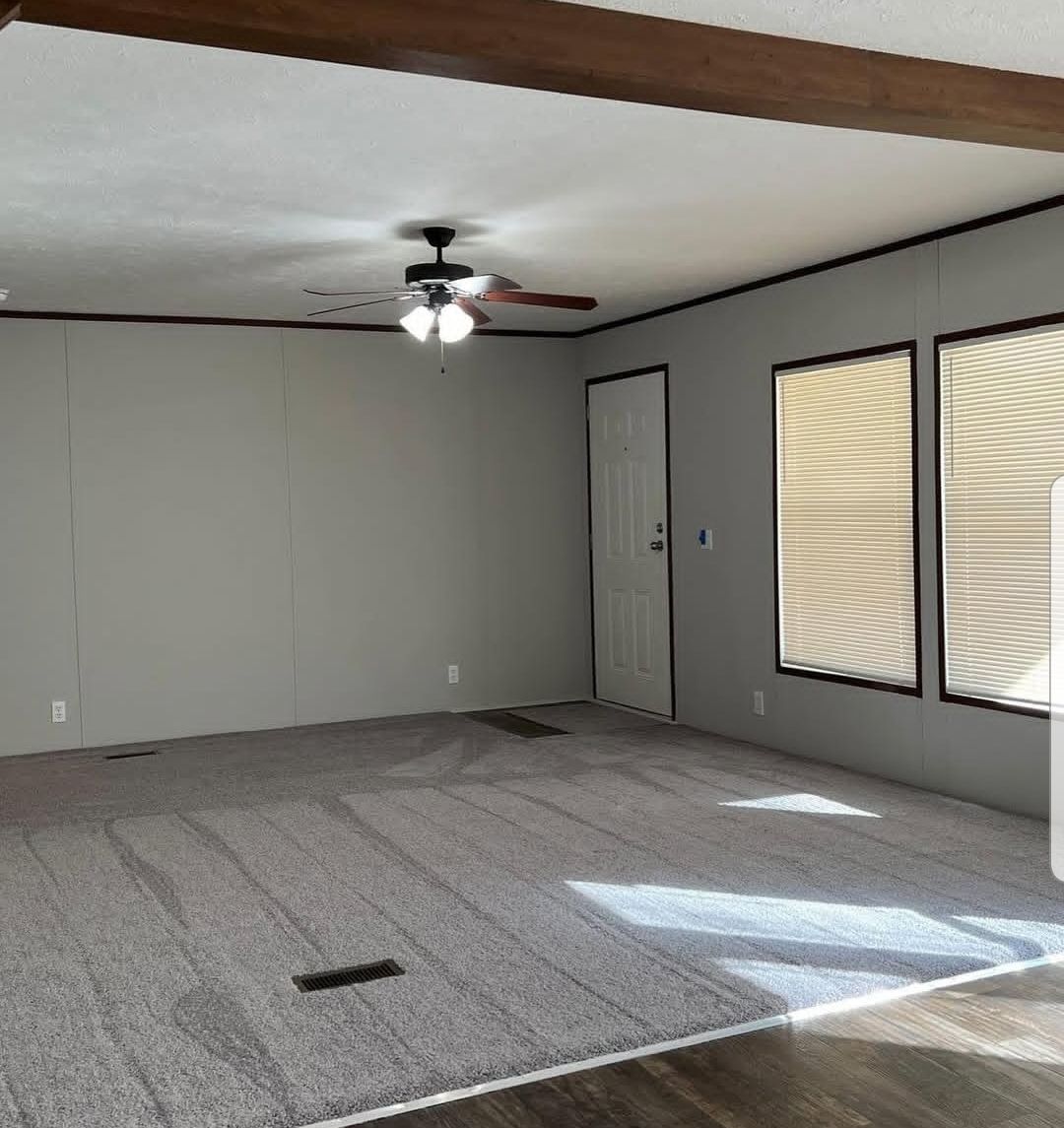 A spacious, empty room with gray carpet, neutral walls, and a wooden ceiling beam. Sunlight streams through blinds on two large windows, creating a calm ambiance.