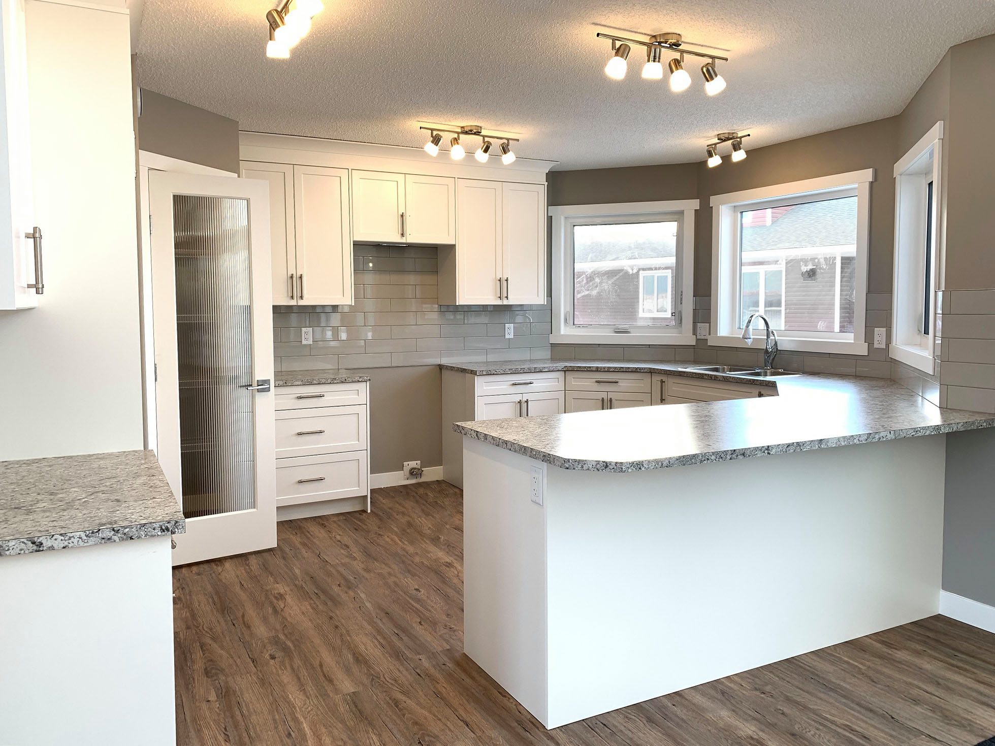 Modern kitchen with white cabinets, gray countertops, and wood flooring. Bright lighting and large windows create an inviting and airy feel.