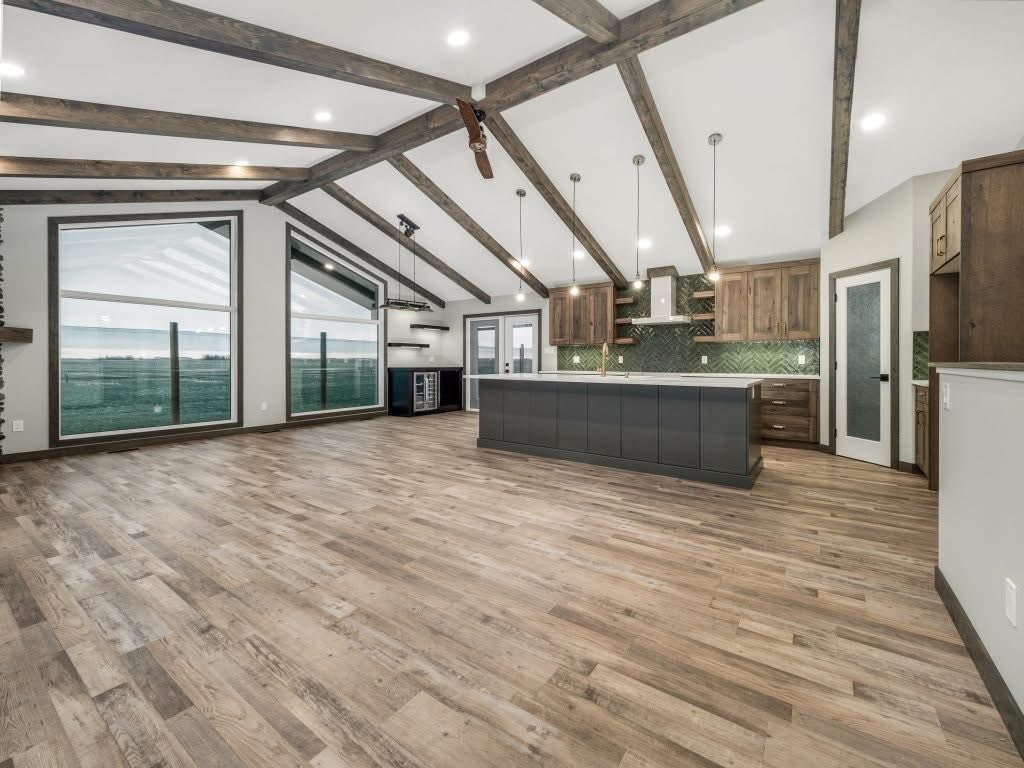 Spacious modern kitchen with vaulted ceiling and wood beams. Features large windows, wood floor, gray island, and light wood cabinets. Bright and airy atmosphere.