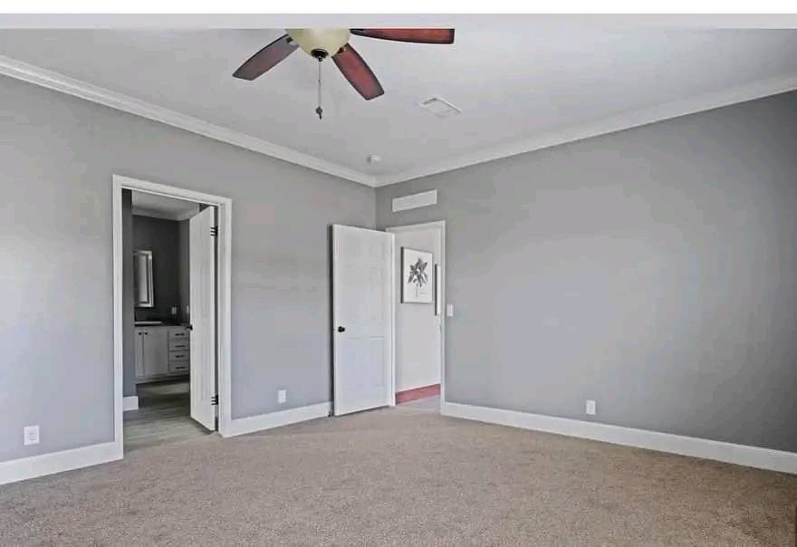 A spacious, empty room with gray walls and carpet. A ceiling fan with wooden blades hangs above. An open door leads to a bathroom with visible cabinets.