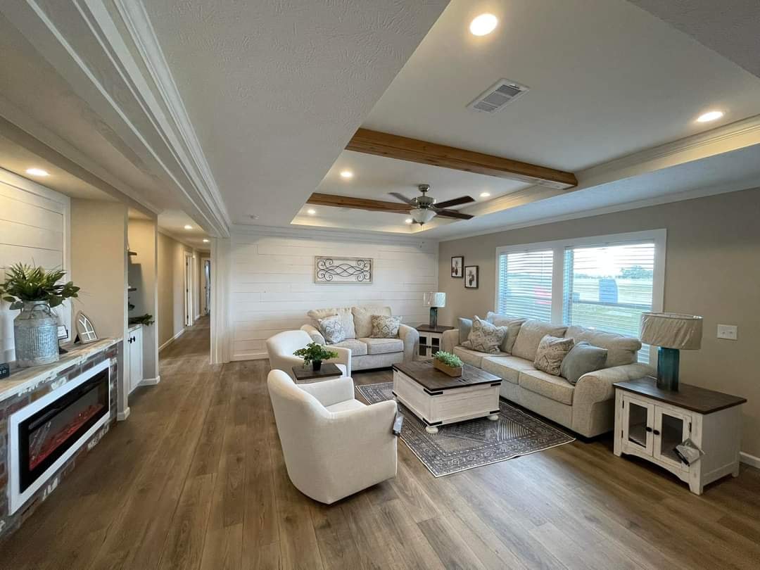 Cozy living room with beige sofas, armchairs, and a wooden coffee table on a rug. Decorative beams on ceiling, a fan, and large windows create a welcoming space.