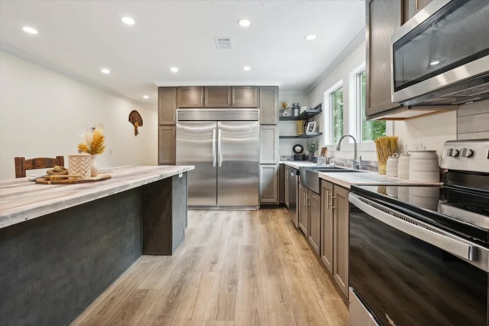 Modern kitchen with wooden flooring, sleek gray cabinets, and stainless steel appliances. A central island with decor and large windows add brightness.