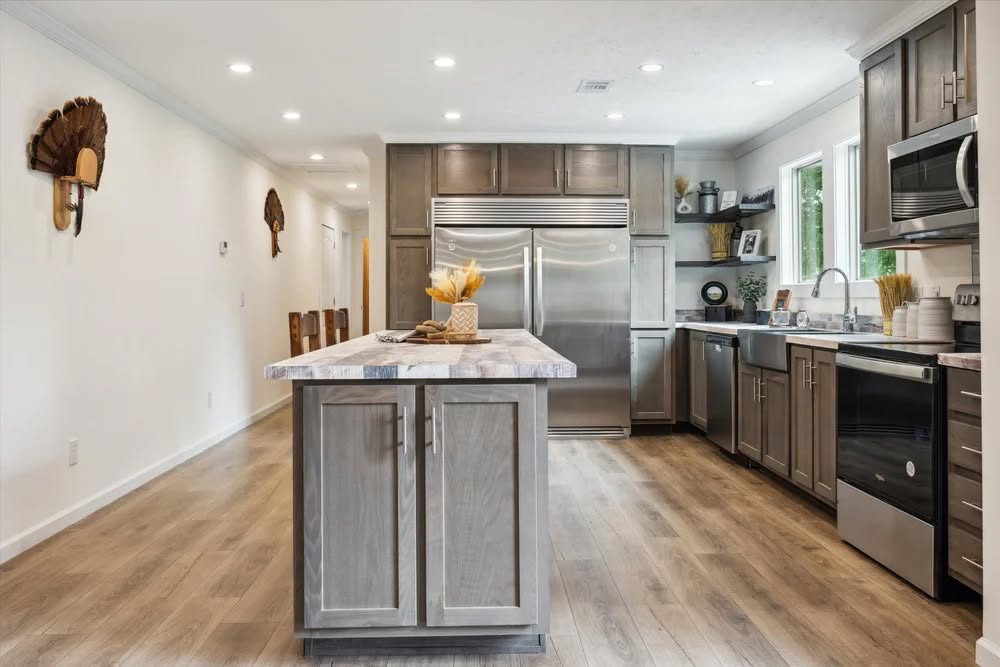 Modern kitchen with gray cabinetry, stainless steel appliances, and a central island topped with a plant arrangement. Wooden floor and ample natural light enhance the space.