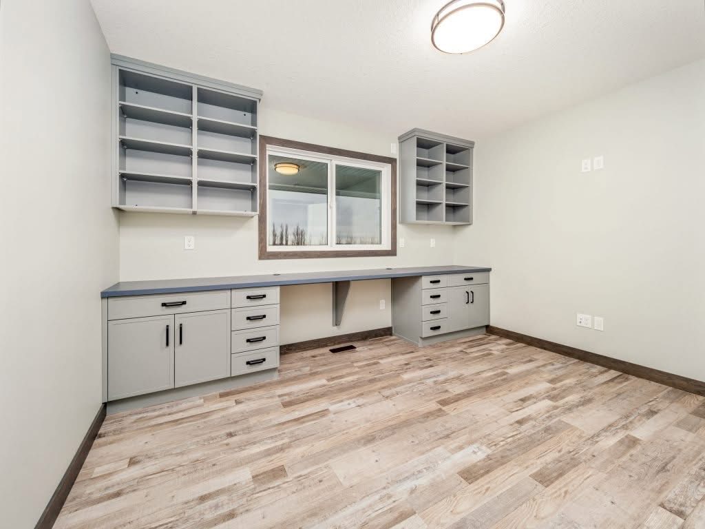 Spacious home office with light wood floors, gray cabinetry, and a large built-in desk under a window. Shelves and drawers offer ample storage.